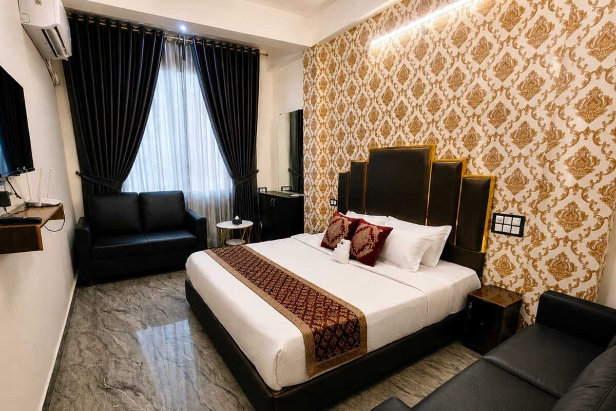 A hotel room with a bed, a sofa, a desk with a lamp, and a wall-mounted air conditioner.