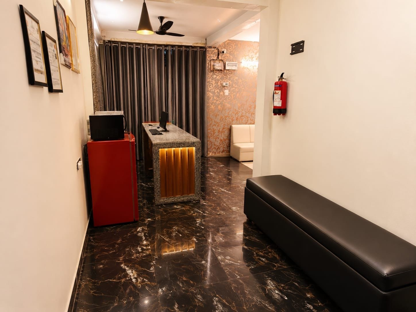 A hotel lobby with a reception desk, a red refrigerator, a bench, a fire extinguisher, and a patterned wall.