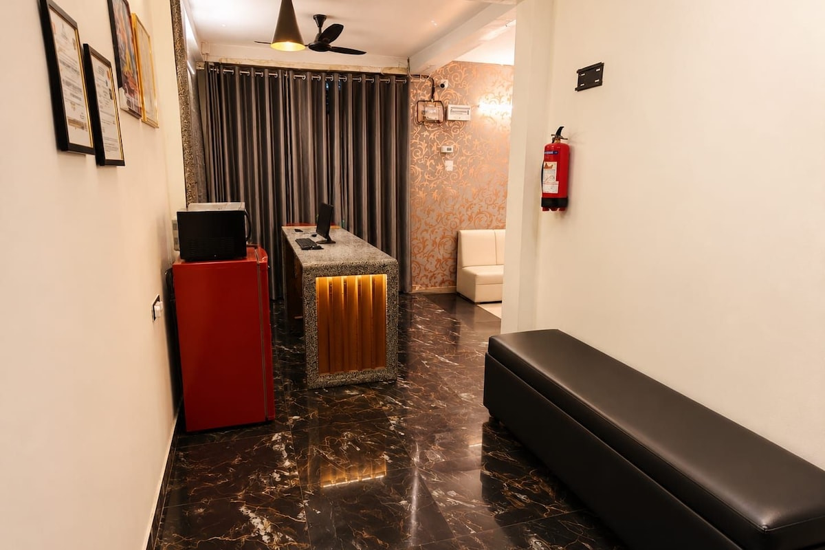 A hotel lobby with a reception desk, a red refrigerator, a black bench, a fire extinguisher, and a patterned wall.