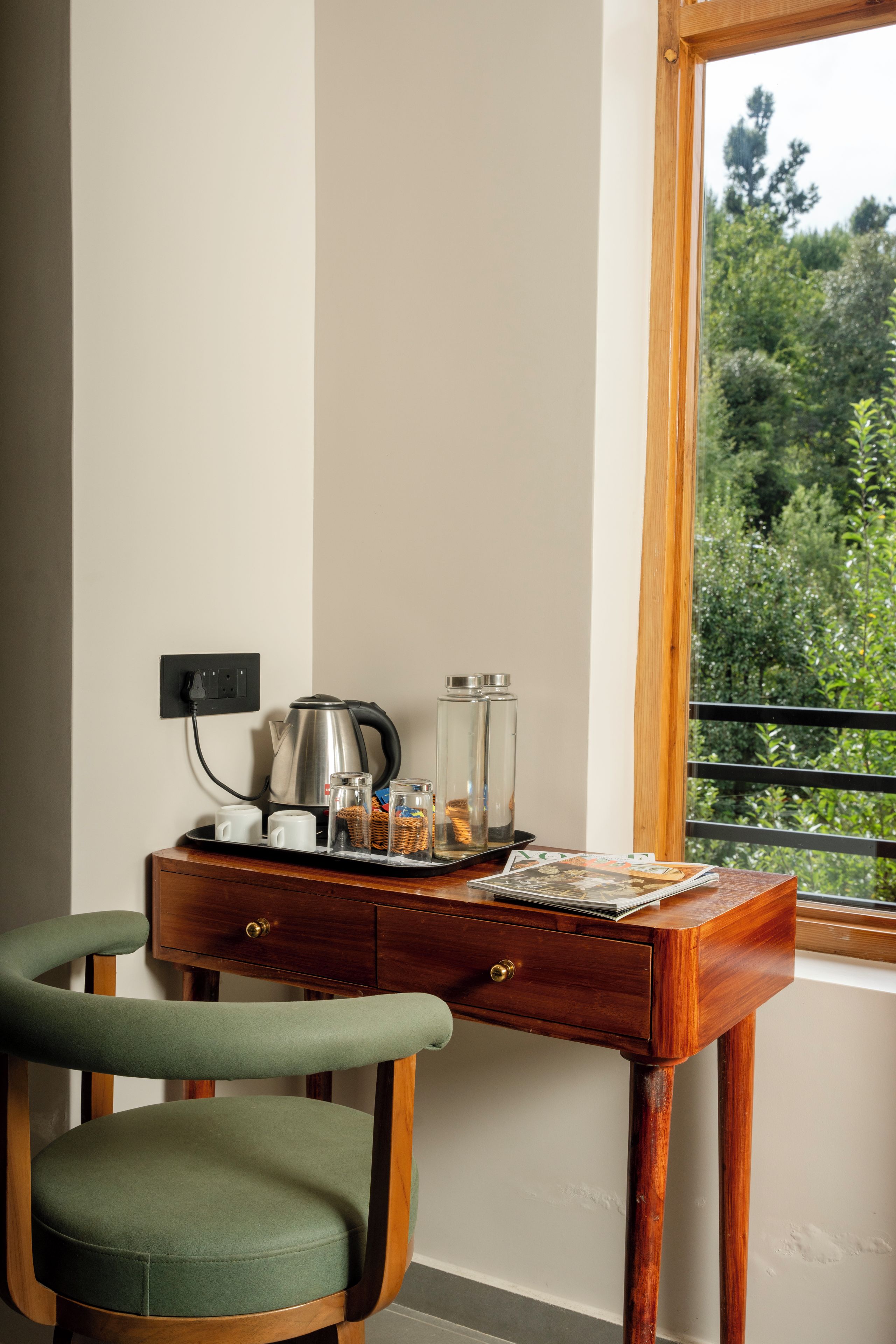 Escritorio de madera con una tetera, tazas y recipientes de vidrio, junto a una silla tapizada de verde, con vista a follaje.