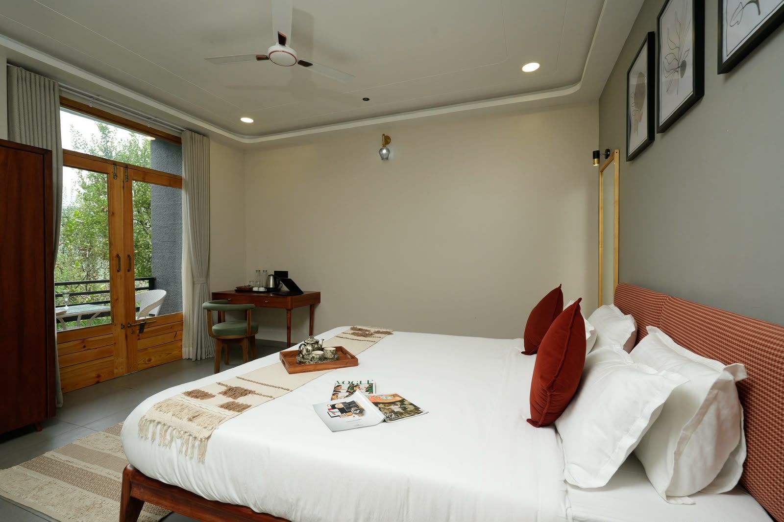 Un dormitorio con cama, escritorio y silla. Hay una ventana con cortinas, un ventilador de techo y una obra de arte enmarcada en la pared.