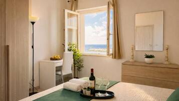Habitación de hotel con cama, escritorio, silla, ventana con vista al mar, una botella de vino y dos copas sobre una bandeja.