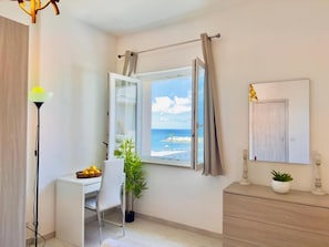 Una habitación con escritorio, silla, planta en maceta y vista al mar a través de una ventana abierta.