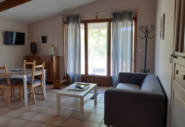 A living room with a sofa, a dining table with chairs, a TV mounted on the wall, and a coat rack.