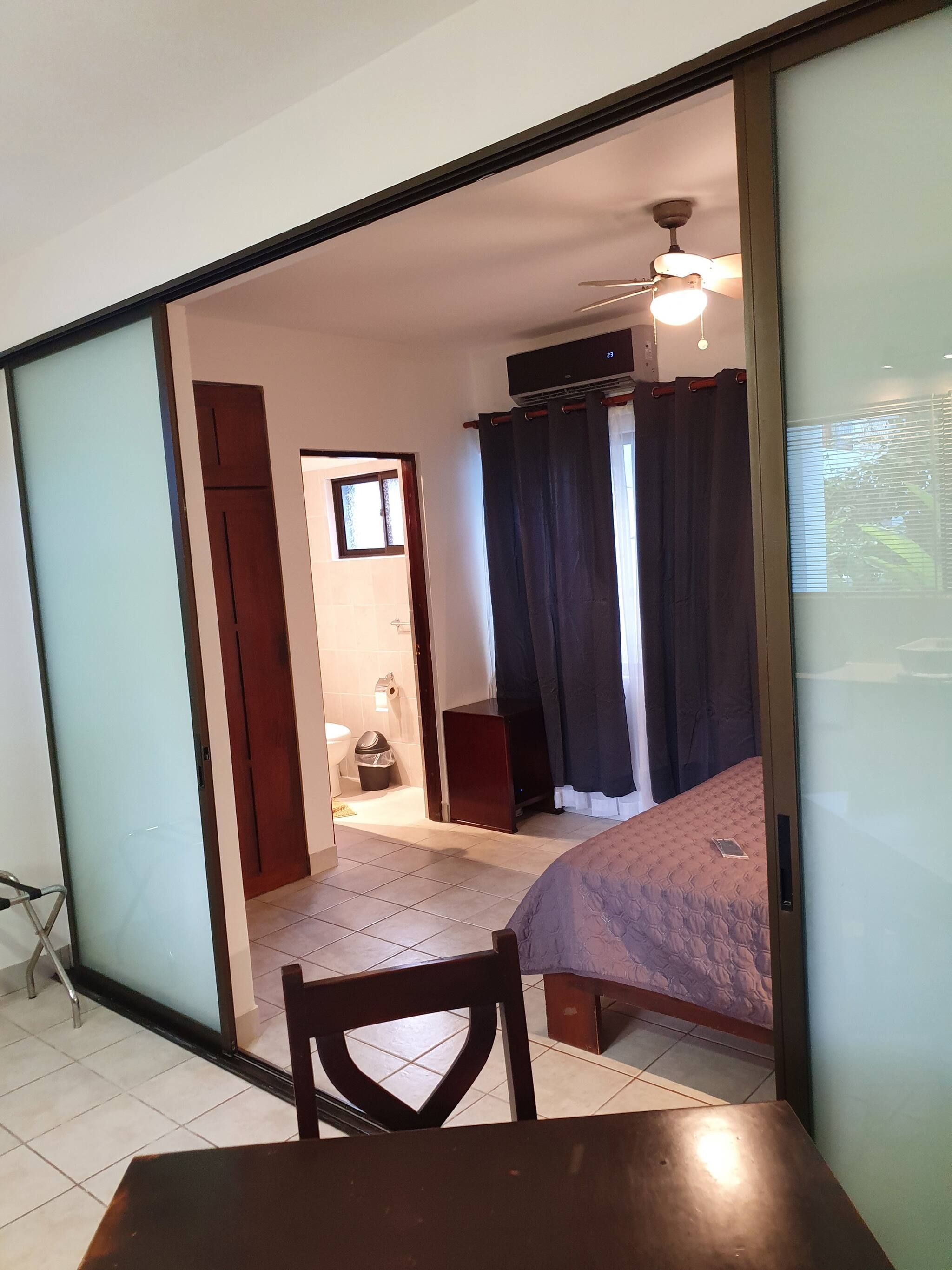 Una habitación con una cama, una silla, una mesa, un ventilador de techo y un ventanal.