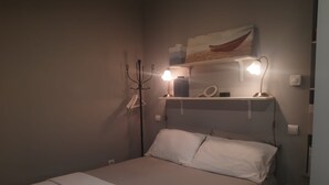 A single bed with white bedding, a nightstand with a lamp, a wall-mounted shelf with a mirror and decorative items, and a wall-mounted light fixture. - Holiday Home 'Estudio Minimalista En Galizano' with Private Terrace, Private Garden and Wi-Fi (Cantabria)
