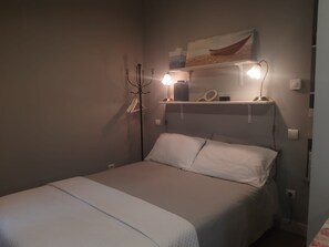 Un lit simple avec une literie blanche, une table de chevet avec une lampe, une étagère murale avec un miroir et des objets décoratifs, et un luminaire fixé au mur.