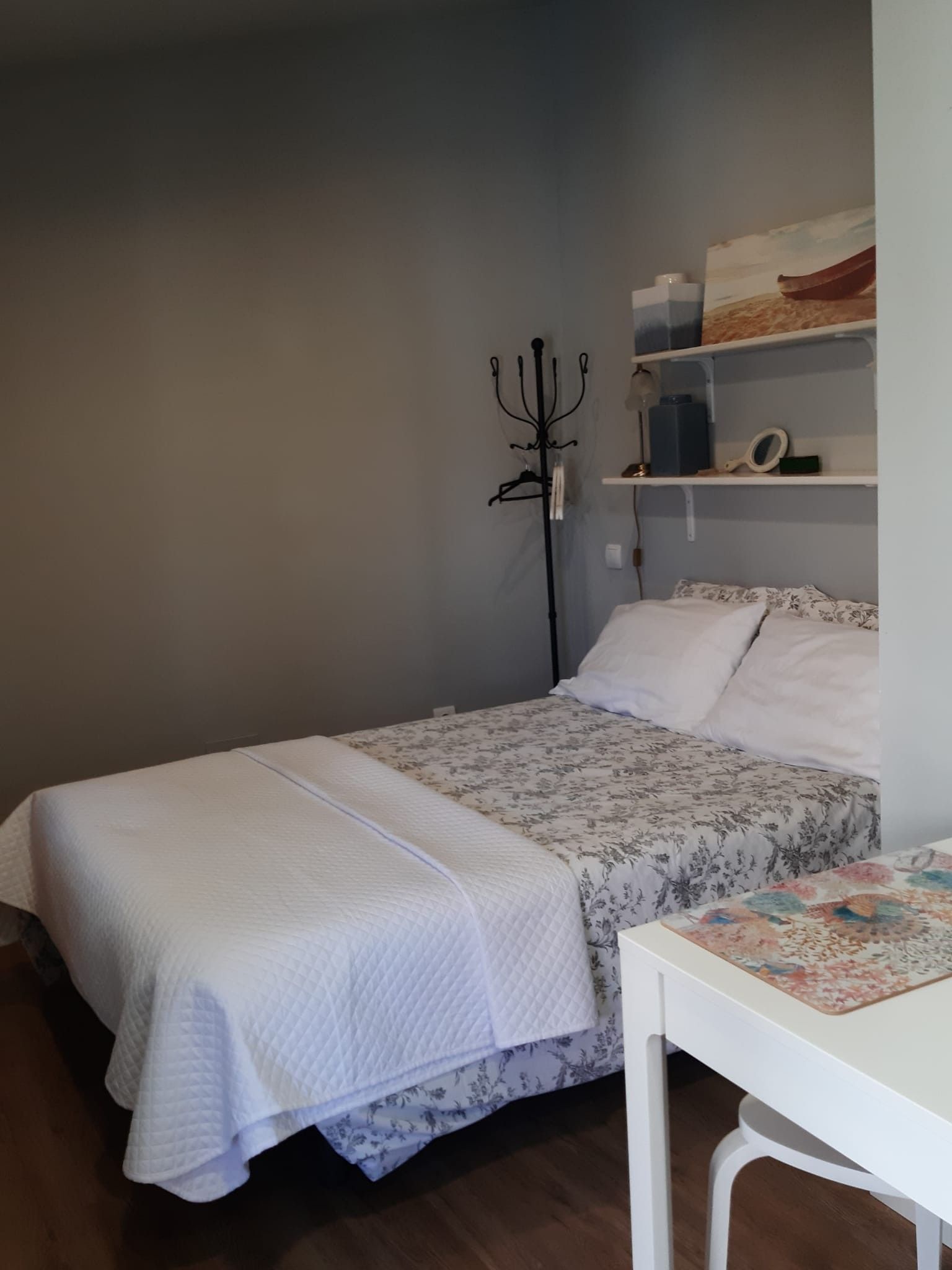 A bedroom with a bed, a bedside table, a shelf with decorative items, and a wall-mounted lamp.