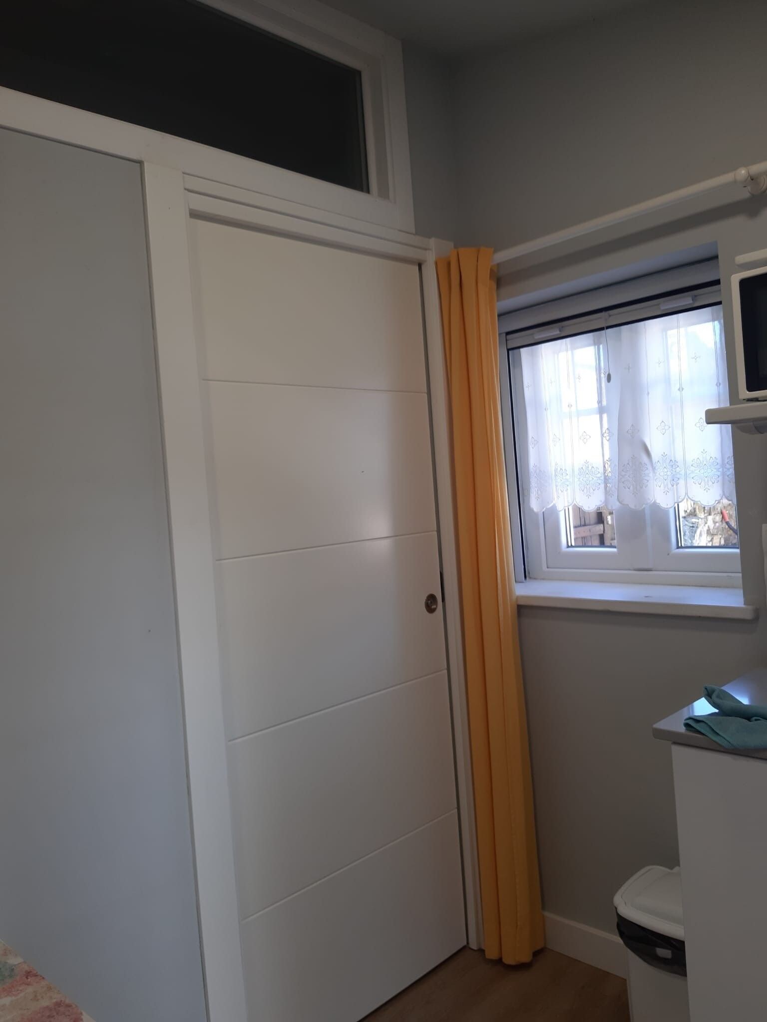 A room with a white wardrobe, a window with a curtain, and a small table with a trash bin.