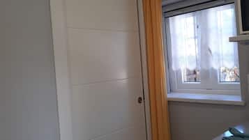 A room with a white wardrobe, a window with a curtain, and a small table with a trash bin.