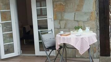 Una habitación con pared de piedra, una mesa con mantel blanco, una silla y un ventanal con persianas blancas.