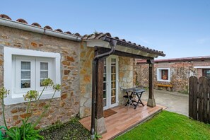 A stone house with a tiled roof, a covered patio with a table and chairs, and a small garden area.