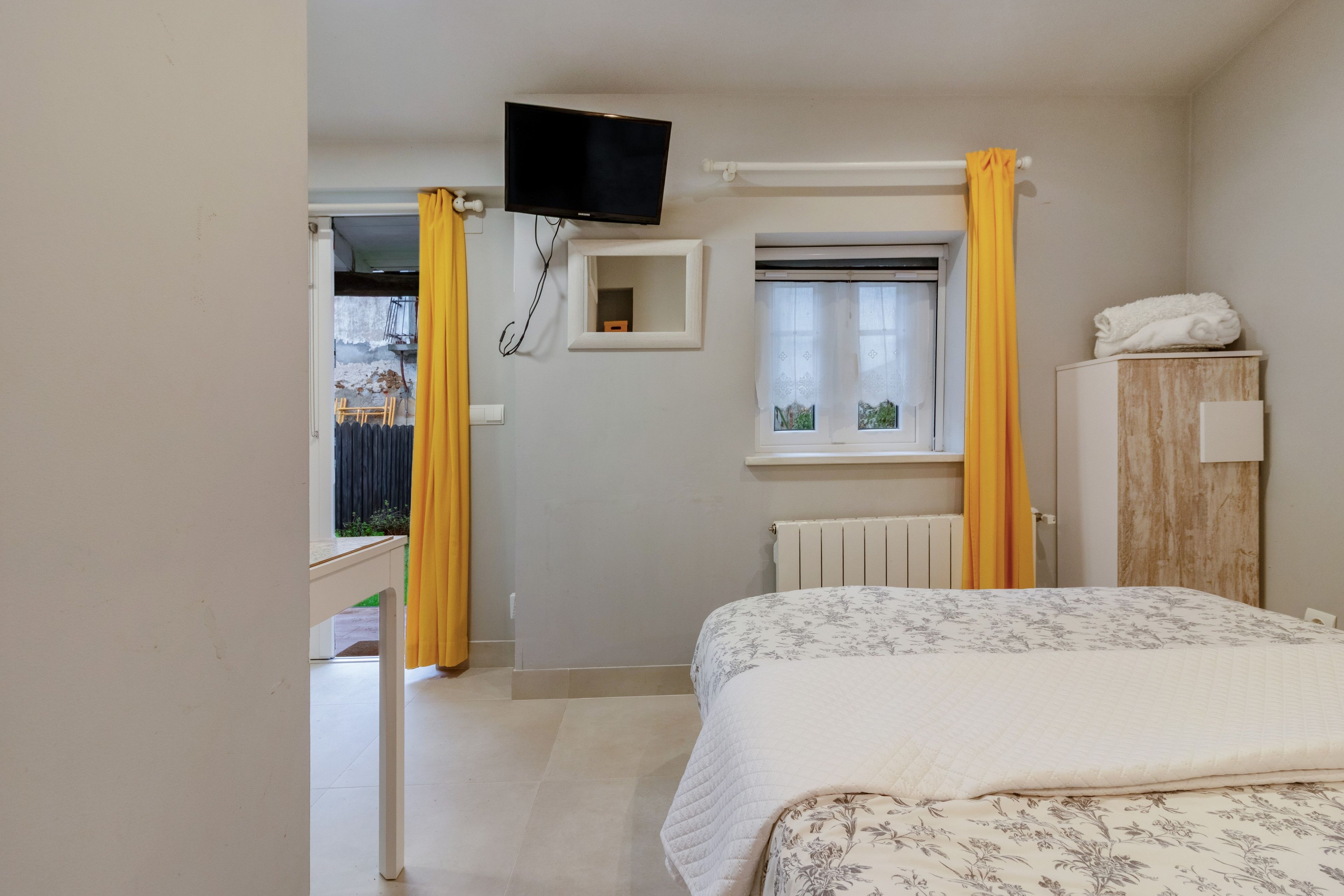 Un dormitorio con cama, televisor montado en la pared, ventana con cortinas y radiador.