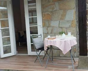 Une pièce avec un mur en pierre, une table recouverte d’une nappe blanche, une chaise et une porte donnant sur une autre pièce.