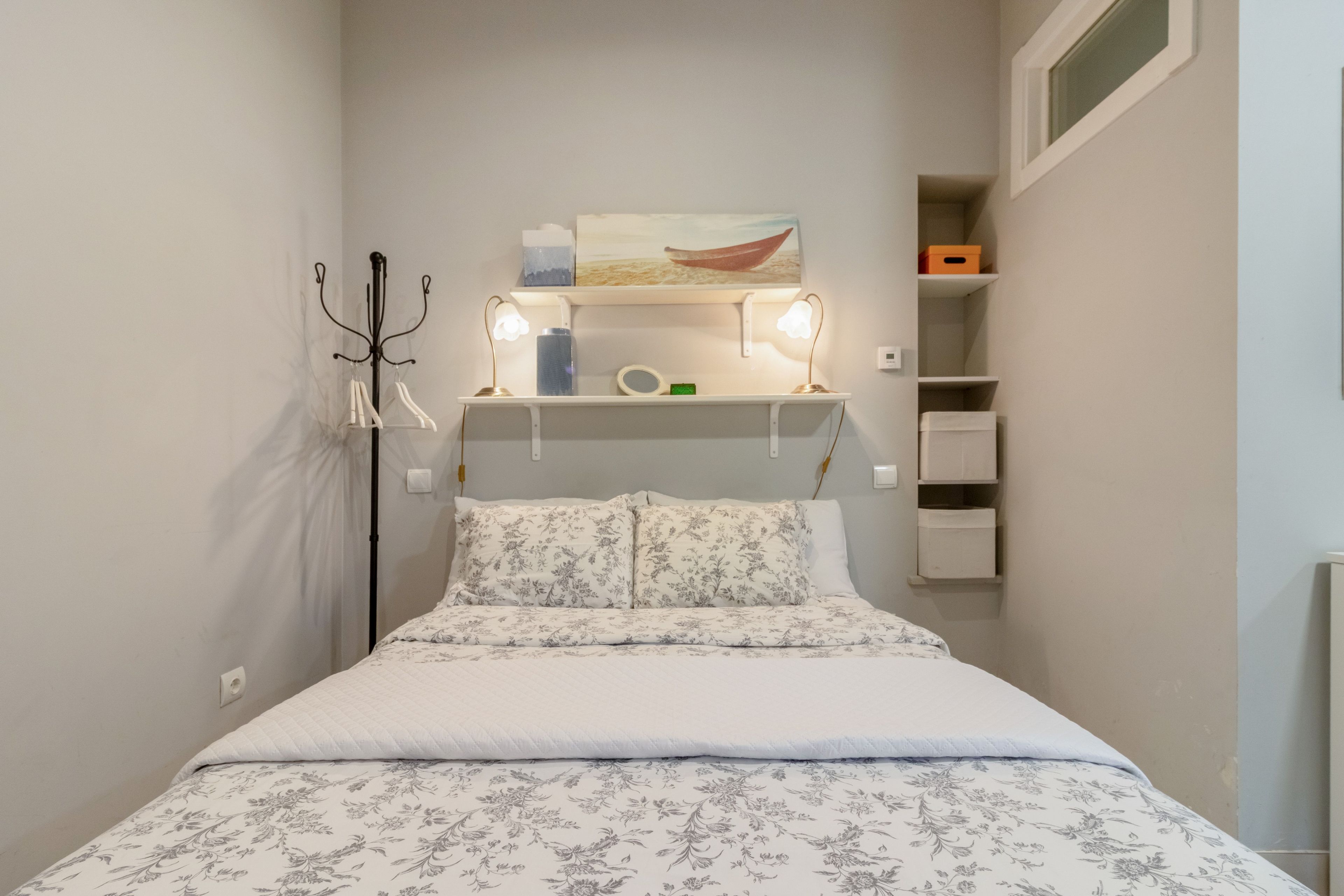 Un dormitorio con una cama, una mesita de noche, una lámpara, un estante con objetos y un cuadro en la pared.