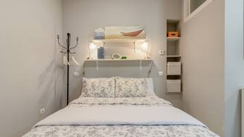 Un dormitorio con una cama, una mesita de noche, una lámpara, un estante con objetos y un cuadro en la pared.