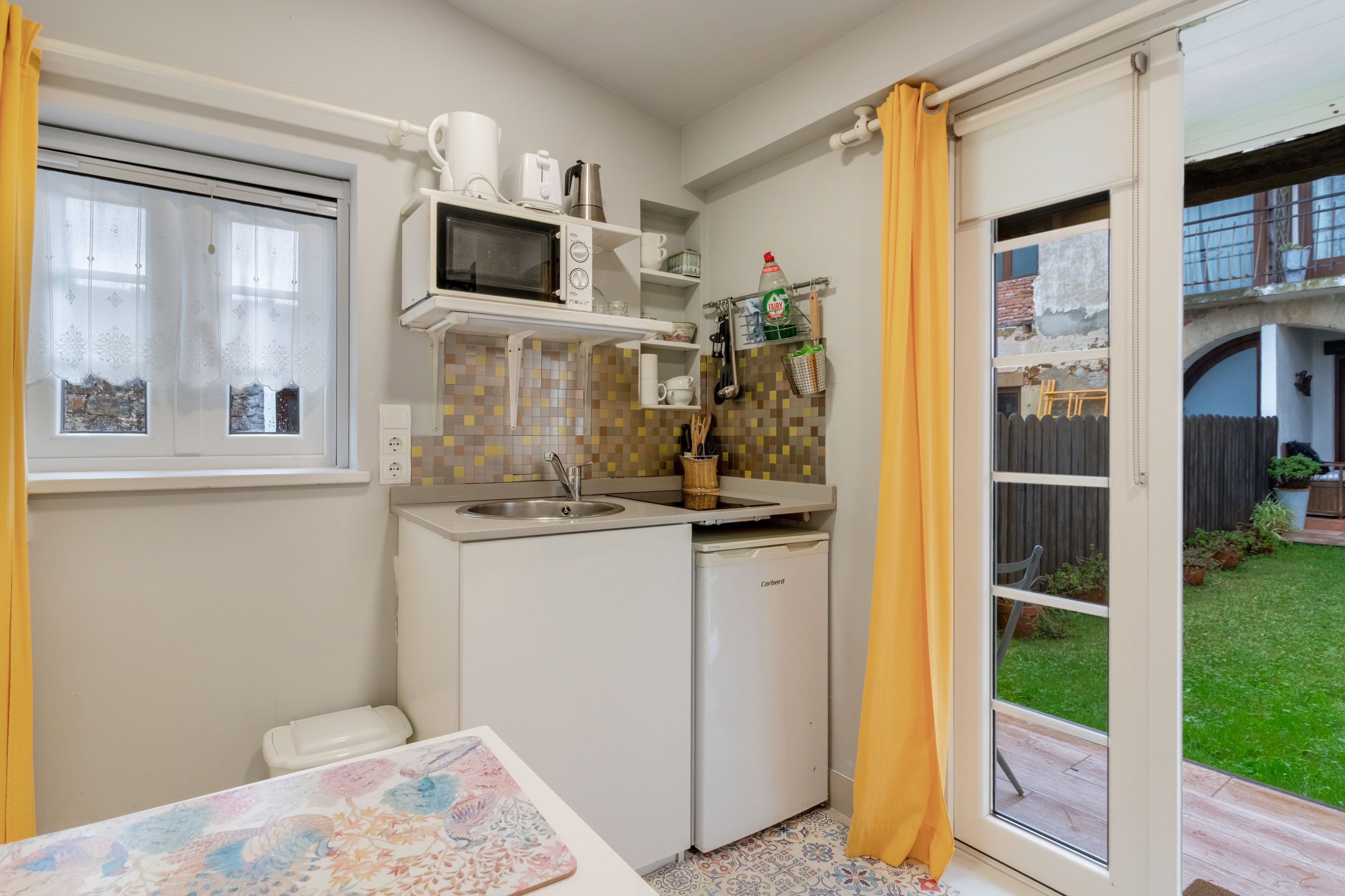 Eine kompakte Küche mit Mikrowelle, Spüle und kleinem Kühlschrank. Ein Fenster mit Vorhängen bietet Blick auf einen Garten.