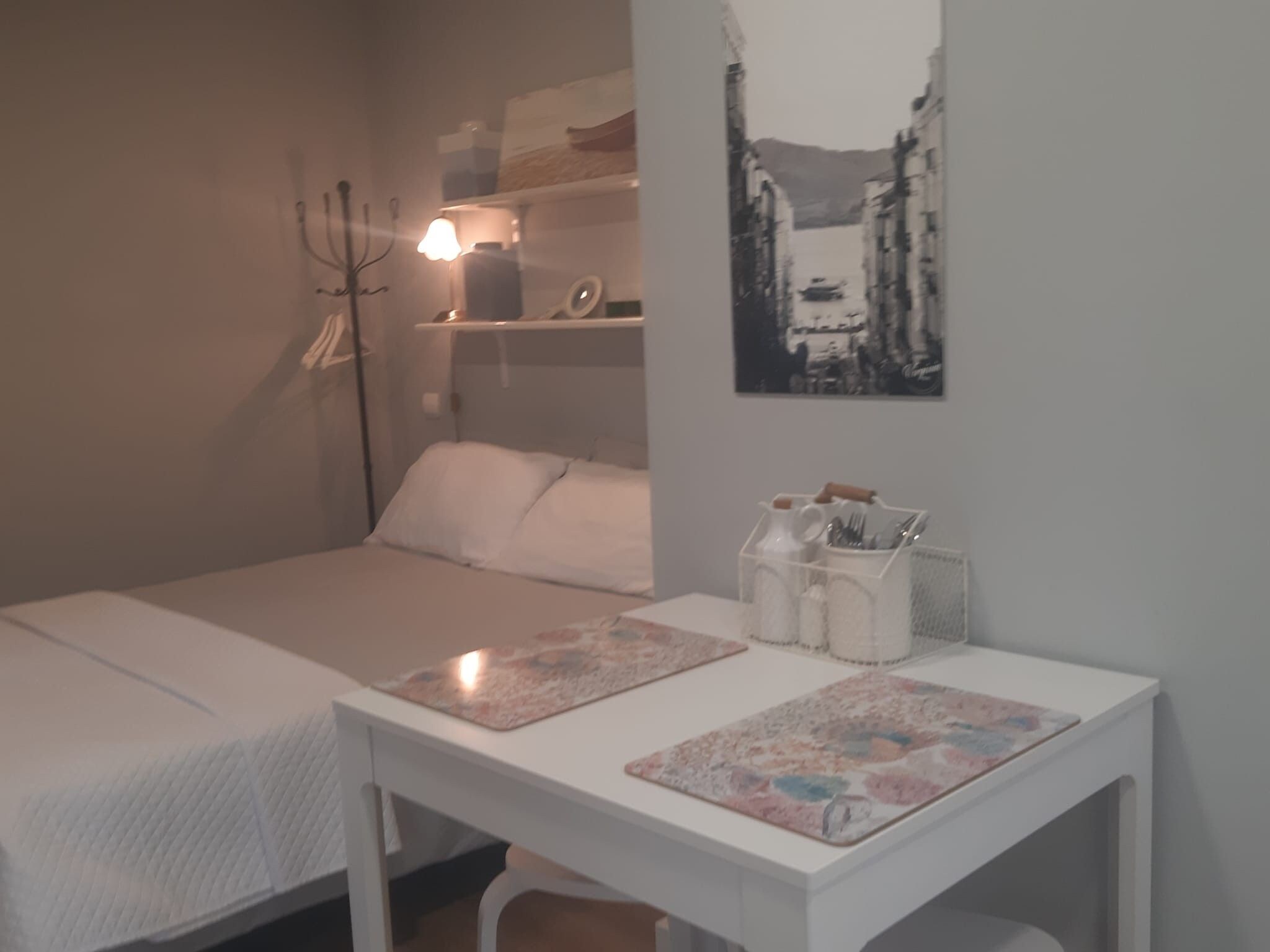 Un dormitorio pequeño y ordenado con una cama individual, una mesita de noche con lámpara, una estantería con objetos personales y un cuadro en la pared.