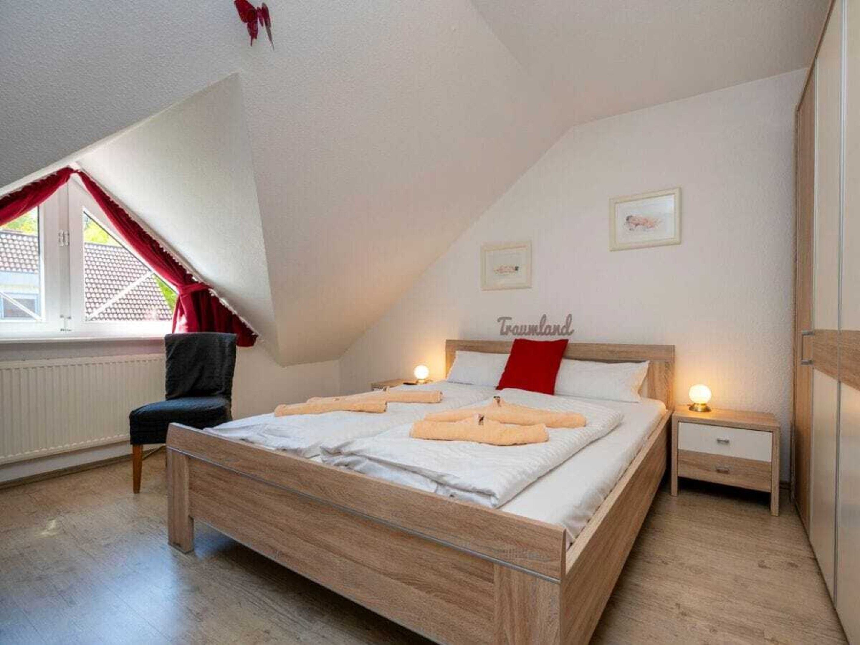 Une chambre avec un plafond en pente, un lit recouvert de draps blancs, un oreiller rouge, une table de chevet avec une lampe et une fenêtre ornée de rideaux rouges.