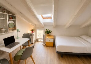 Ein kleines Zimmer mit Schreibtisch, Stuhl, Laptop, Bett und einem Oberlicht.