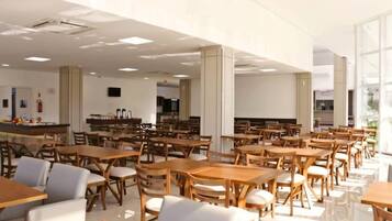 Ampla sala de jantar com várias mesas e cadeiras de madeira, balcão com exposição e grandes janelas que permitem entrada de luz natural.