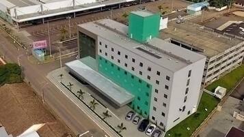 Um edifício de vários andares com telhado plano, uma área verde na lateral e um estacionamento com vários carros.