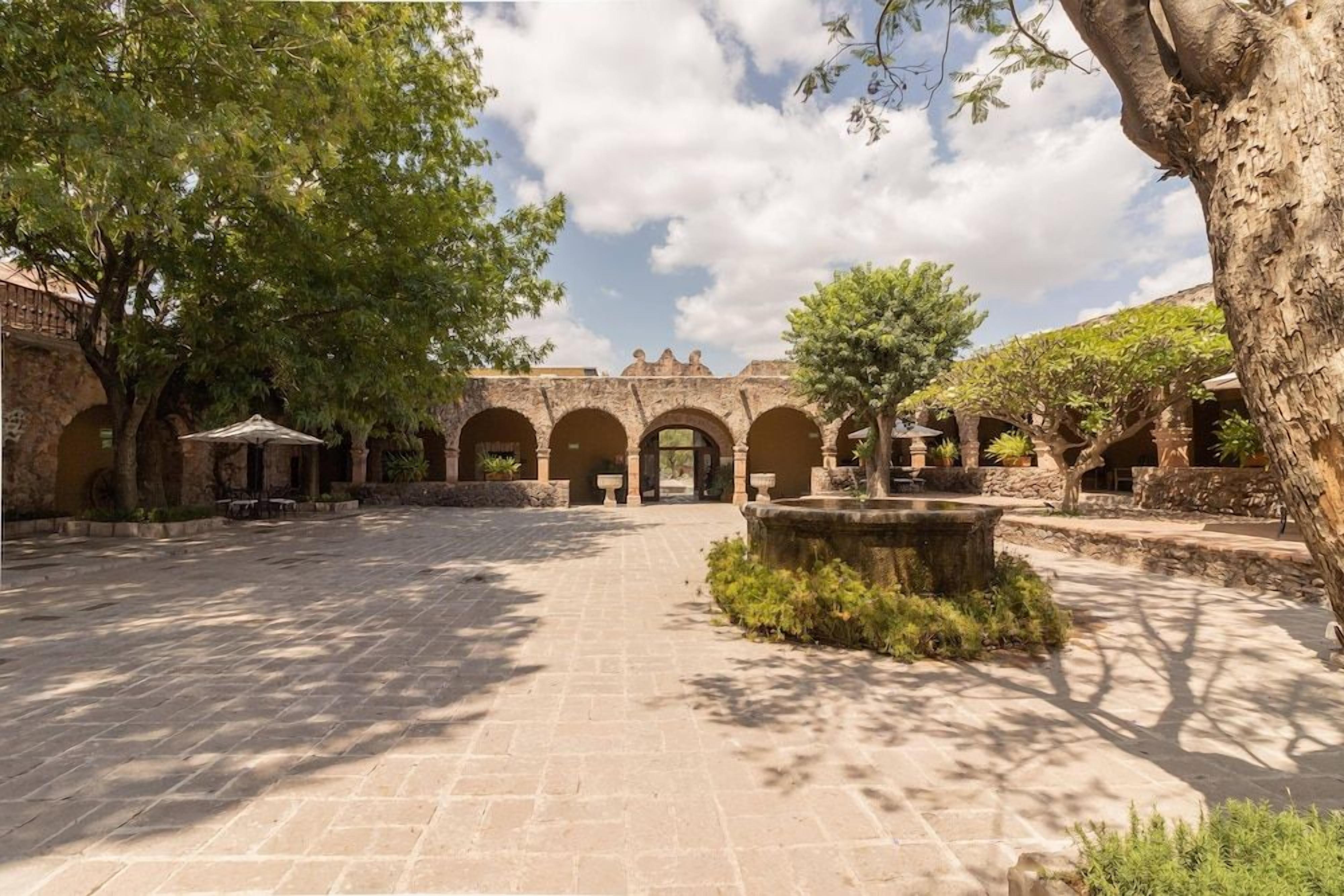 Un patio con suelo de piedra, arcos y árboles que dan sombra.