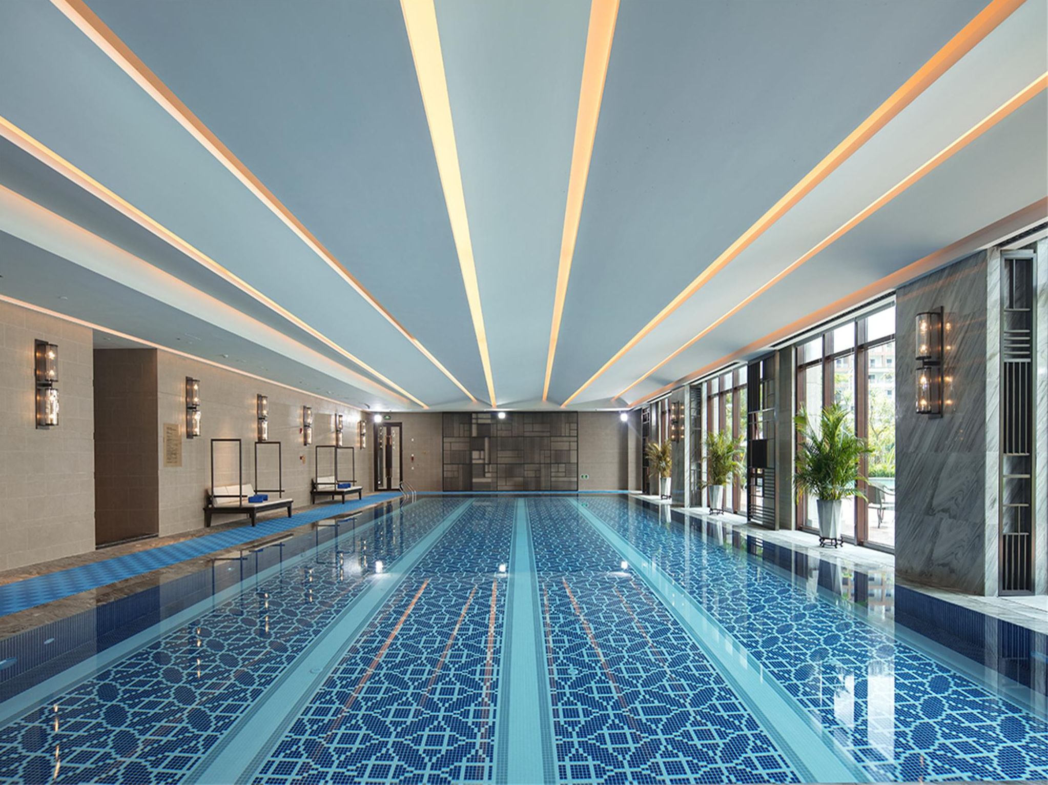 A large indoor swimming pool with a geometric tile pattern, surrounded by walls with recessed lighting and large windows.