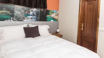 Una cama bien hecha con ropa de cama blanca y una almohada oscura, una puerta de madera a la derecha y una pared con una obra de arte de temática natural sobre la cama.