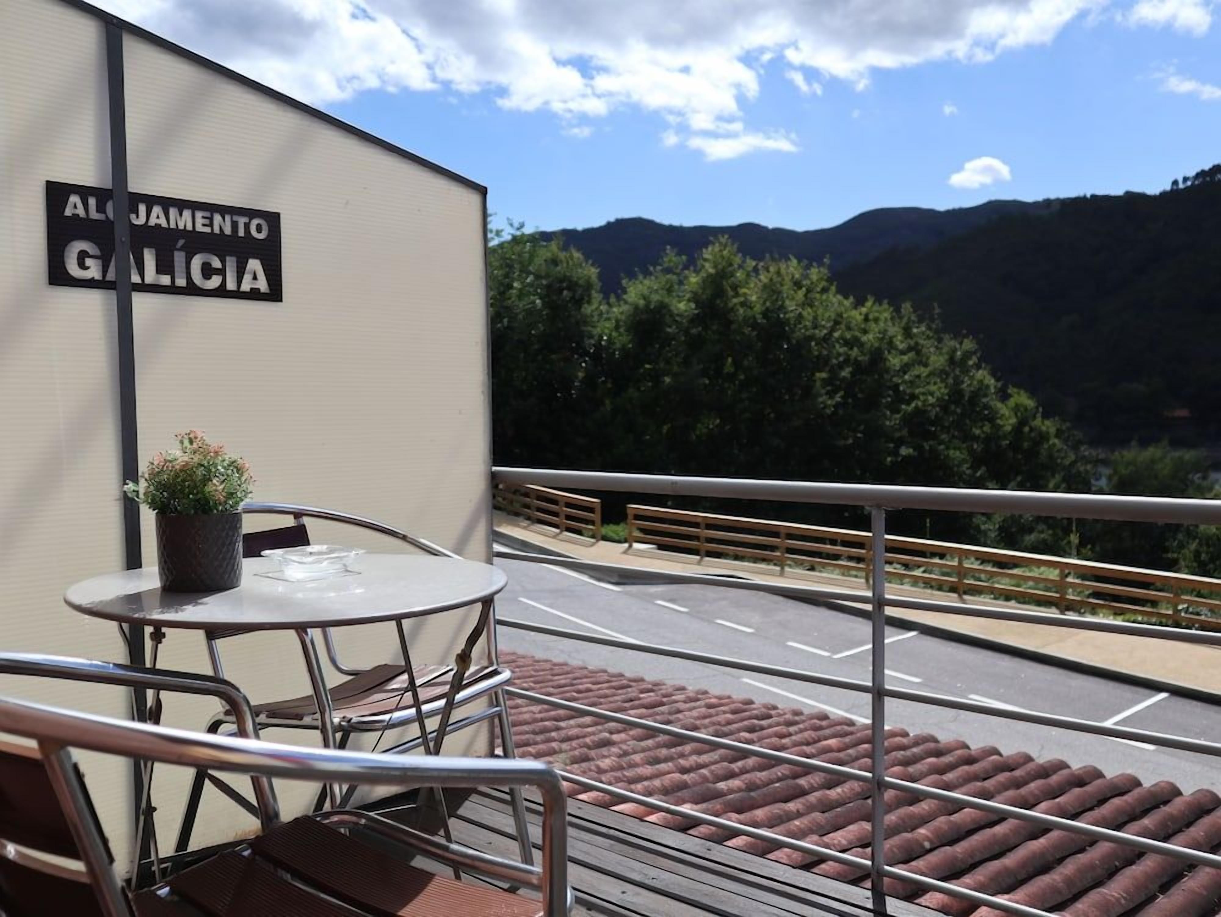 Un balcón con barandilla de metal, una mesa con sillas y una planta en maceta. Al fondo, un cartel que dice 'ALJAMENTO GALICIA' y una vista de un camino con árboles y colinas.