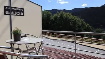 Un balcón con barandilla de metal, una mesa con sillas y una planta en maceta. Al fondo, un cartel que dice 'ALJAMENTO GALICIA' y una vista de un camino con árboles y colinas.