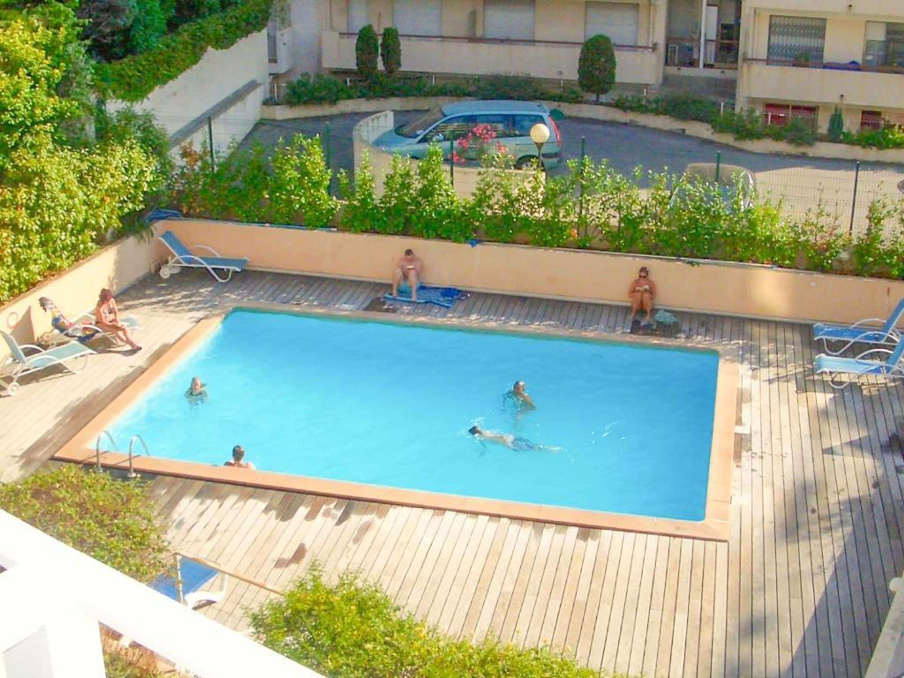 A rectangular swimming pool with a wooden deck, surrounded by lounge chairs, and a view of a residential area with buildings and a parked car.