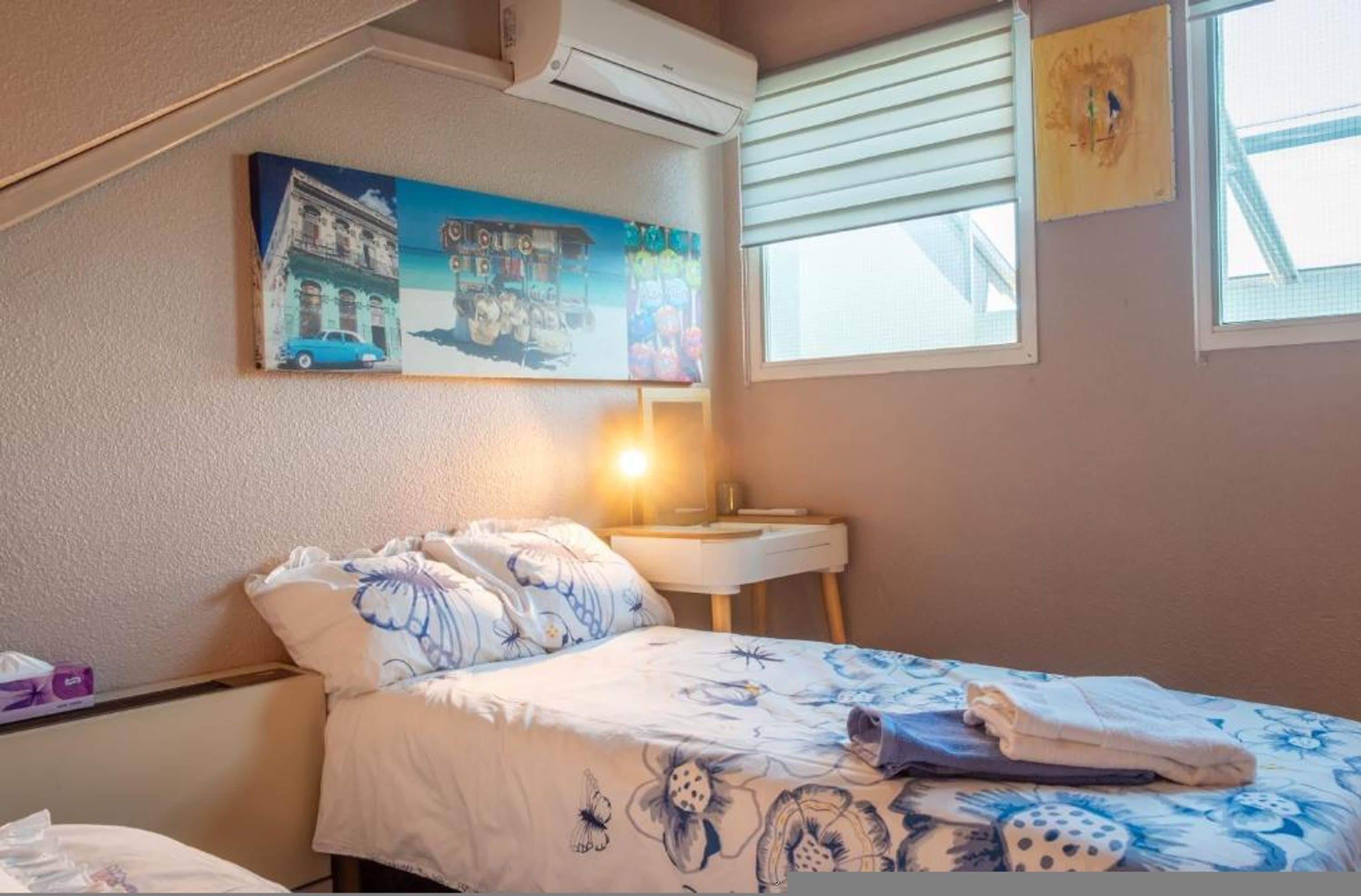 Une chambre à coucher comprenant un lit, une table de chevet, une lampe, une fenêtre avec des stores et un climatiseur fixé au mur.