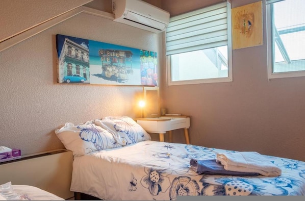 A bedroom with a bed, a nightstand, a lamp, a window with blinds, and a wall-mounted air conditioner.