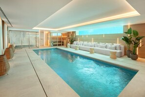 A modern indoor swimming pool area with a white sofa and cushions, a glass door entrance, and potted plants. - Villa In Sierra Blanca - 2398 (Marbella)
