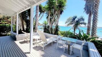 A wooden deck with white chairs and a table, overlooking a beach with palm trees and the sea.