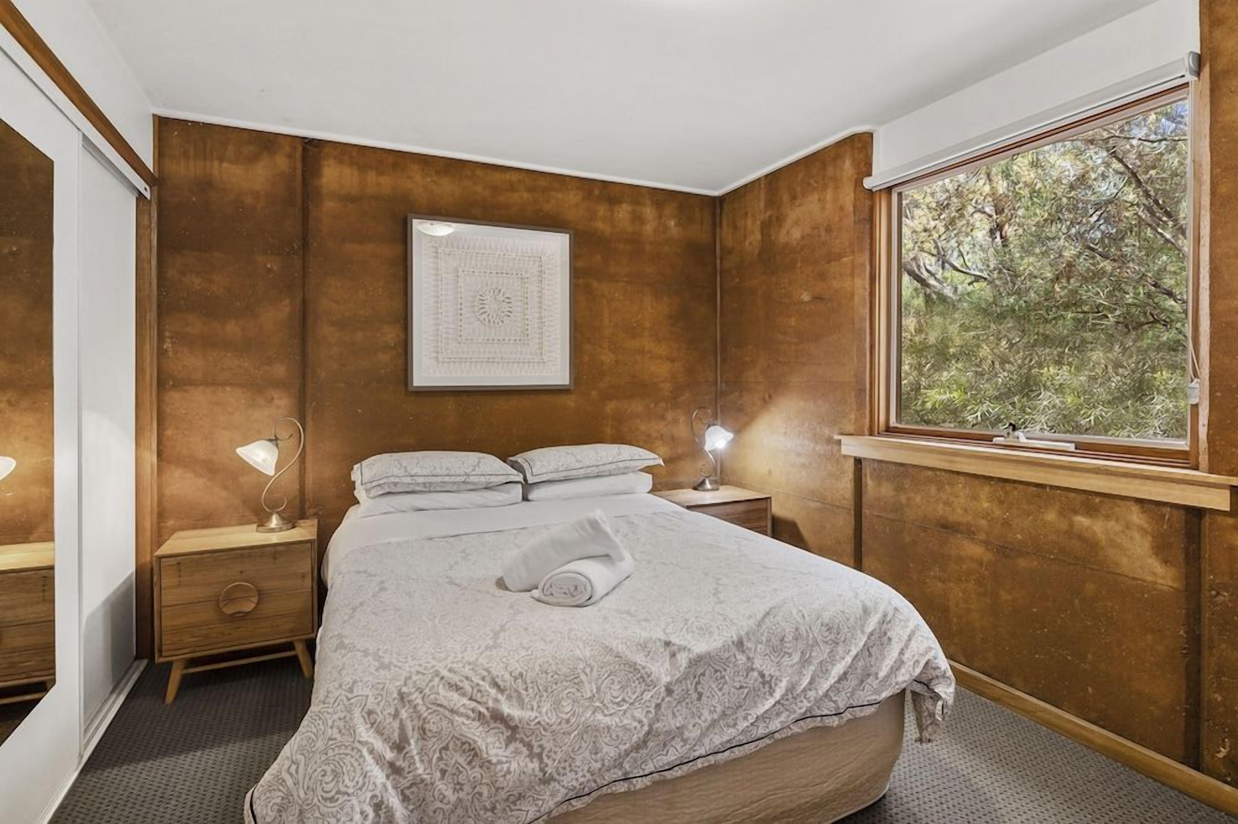 A bedroom with a large bed, two bedside tables, a window with a view of trees, and a framed picture on the wall.