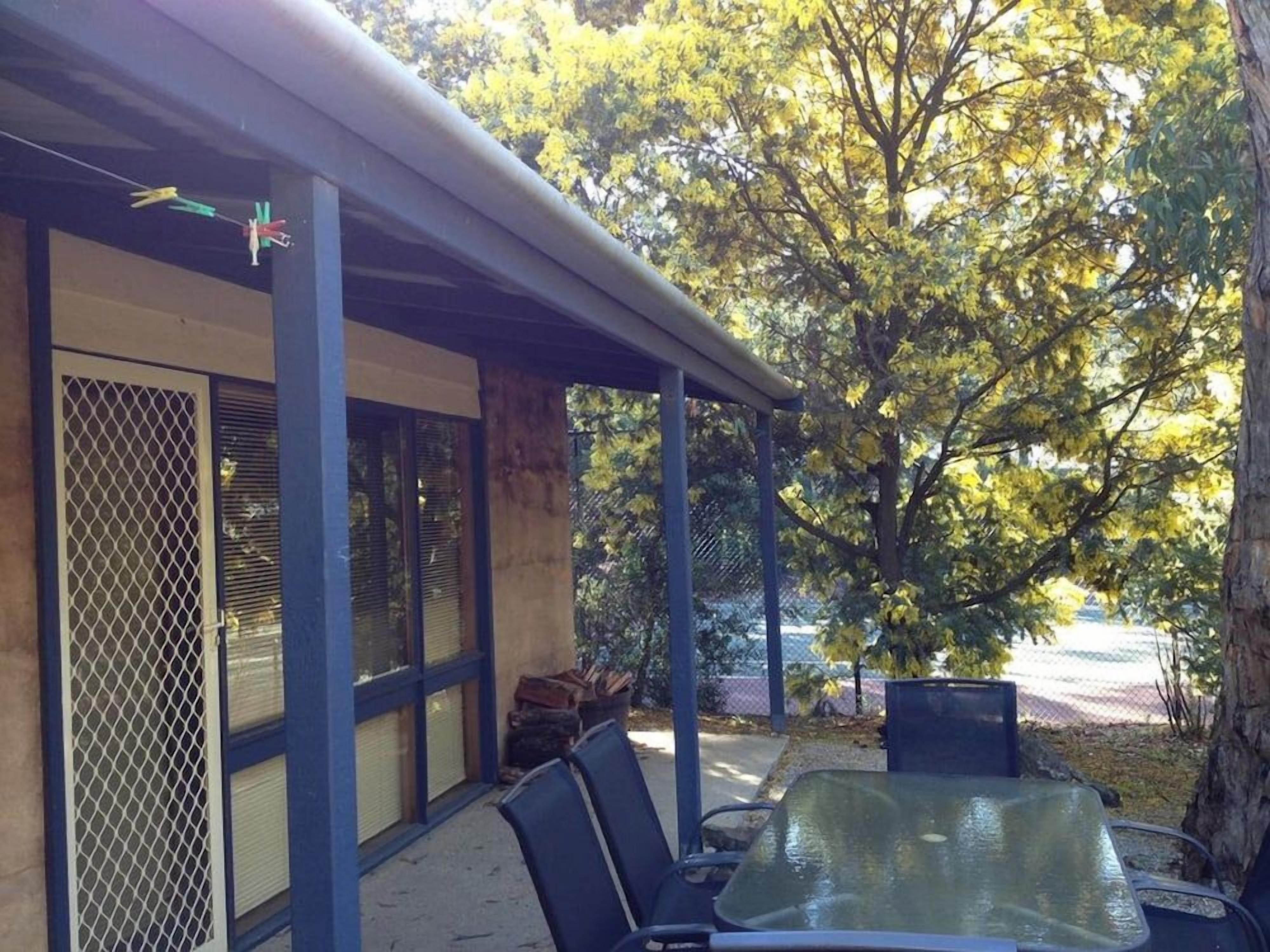 An outdoor seating area with a table and chairs, a covered structure with a mesh window, and trees in the background.