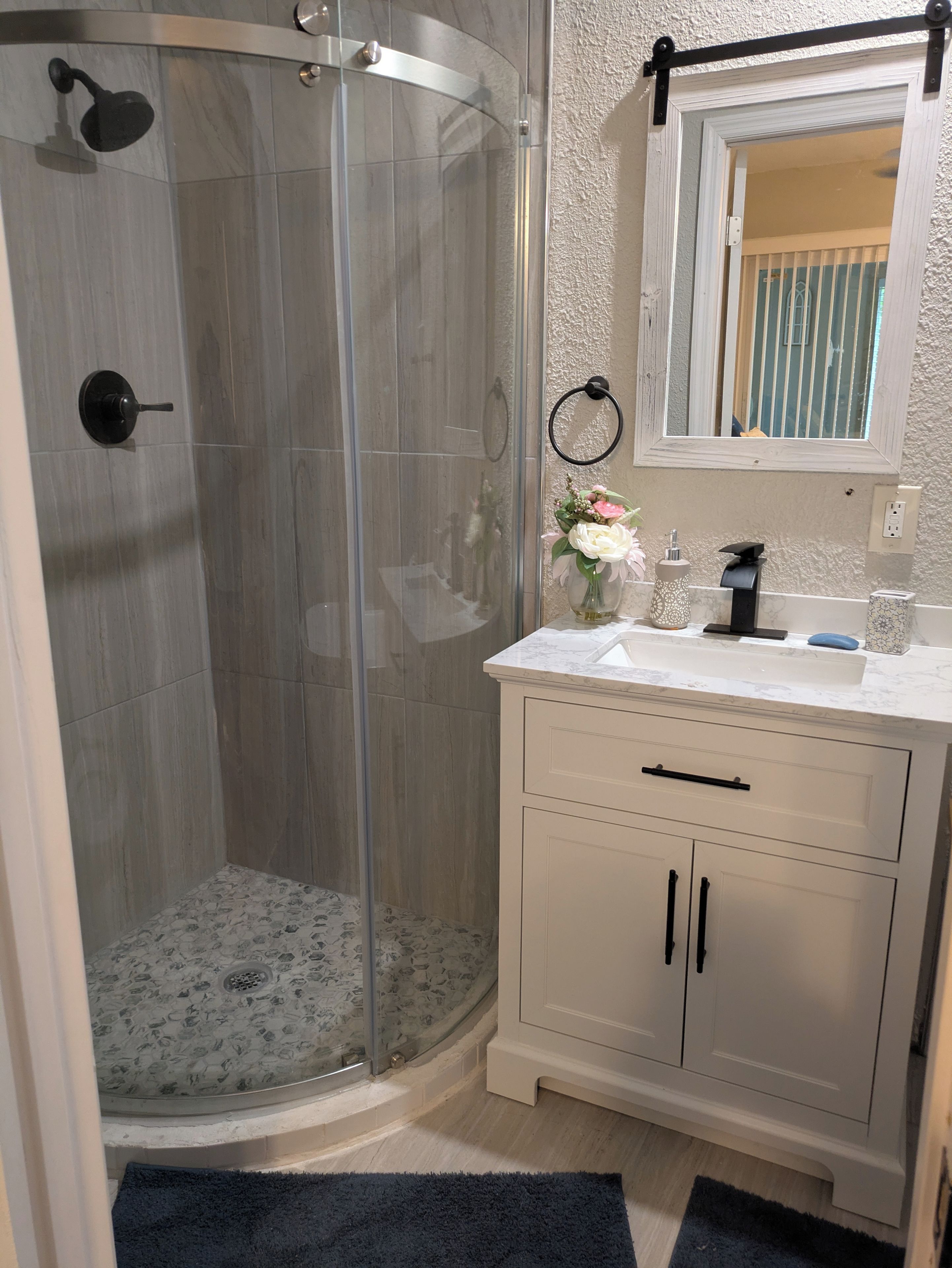 A bathroom with a glass-enclosed shower, a white vanity with a mirror, and a floral arrangement on the countertop.