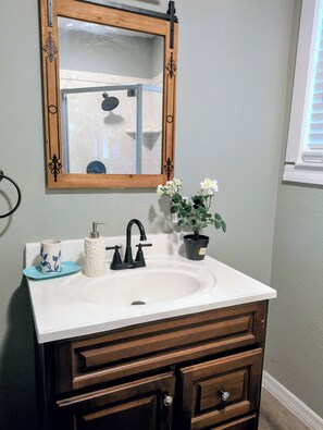 Un baño con un lavamanos blanco, cajones de mueble de madera, un espejo con marco de madera, una planta en maceta y una taza azul sobre el mostrador.