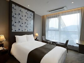 Quarto de hotel com uma cama grande, uma mesa de trabalho, duas cadeiras, uma janela com cortinas e uma parede com padrão geométrico.