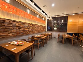 Interior de restaurante com mesas e cadeiras de madeira, parede com padrão e janela com persianas.