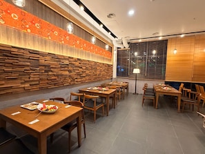 A restaurant interior with wooden tables and chairs, a patterned wall, and a window with blinds.