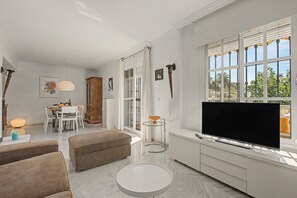 A modern living room with a flat-screen TV, a dining area with a table and chairs, and a kitchen area with a window.