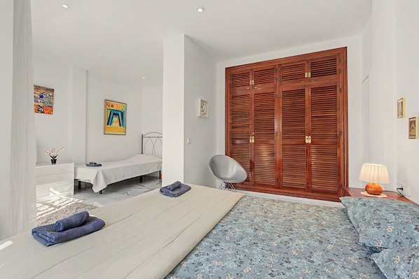 A bedroom with a bed, a bedside table, a chair, a wardrobe, and a small desk with a lamp.