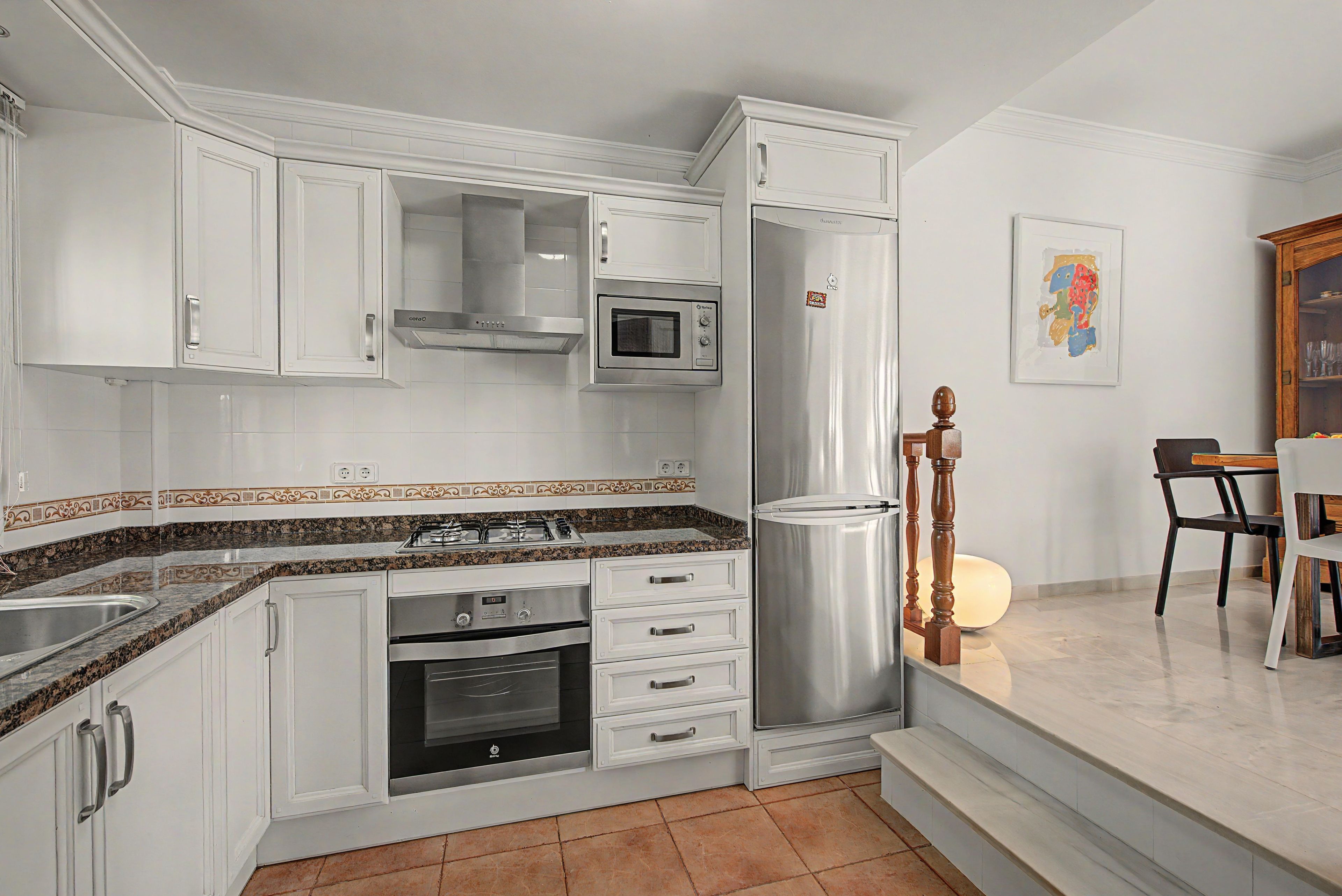 Cocina compacta con armarios blancos, horno integrado, microondas y refrigerador de acero inoxidable.