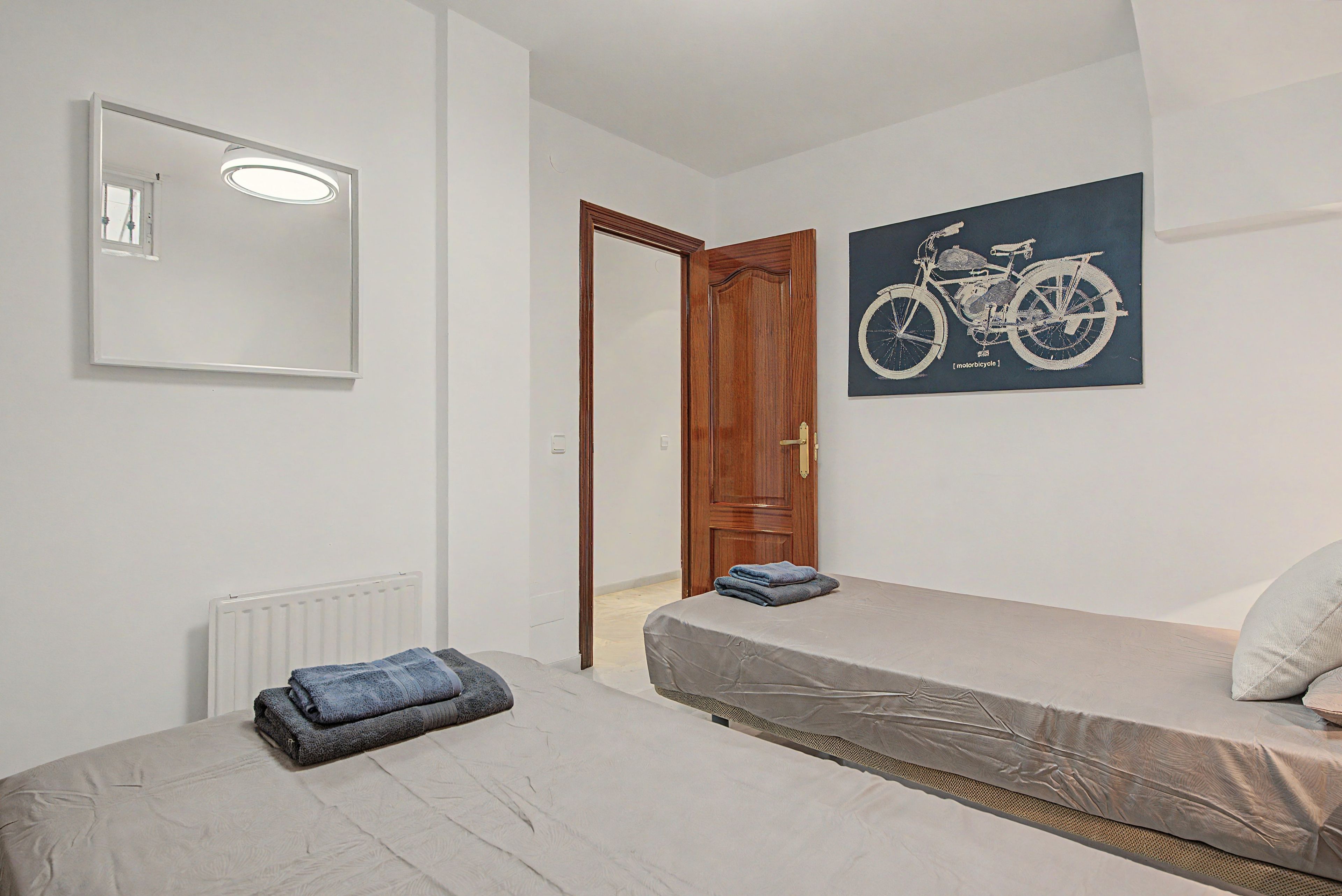 Un dormitorio con cama, un radiador, una puerta, un espejo e ilustración de una bicicleta colgada en la pared.