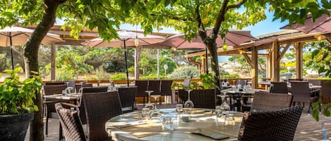 Un área de comedor al aire libre con mesas redondas, sillas de mimbre y sombrillas que dan sombra.