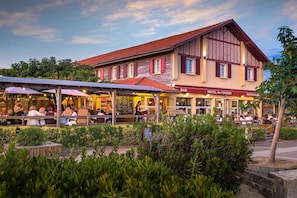 Um edifício com bar e restaurante no andar térreo, área de estar ao ar livre coberta e telhado com telhas vermelhas.
