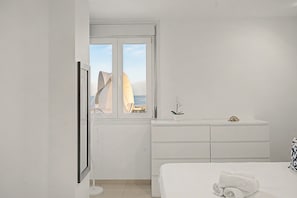 Un dormitorio minimalista con cama, cómoda y ventana con vista a un edificio y al mar.